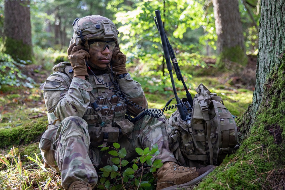 A U.S. Army soldier in full combat gear is seated at the base of a tree in a forested environment, using a radio handset for communication. The soldier is wearing MultiCam (OCP) camouflage uniform, a tactical helmet, and protective eyewear, with face paint applied. A tactical radio with an antenna is visible propped against a backpack beside the soldier.