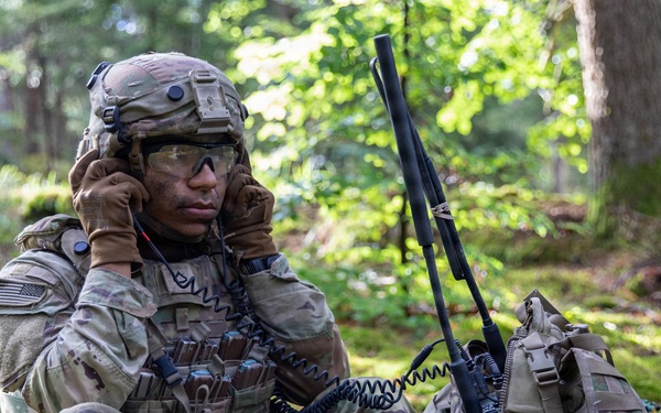 A U.S. Army soldier in full combat gear is seated at the base of a tree in a forested environment, using a radio handset for communication. The soldier is wearing MultiCam (OCP) camouflage uniform, a tactical helmet, and protective eyewear, with face paint applied. A tactical radio with an antenna is visible propped against a backpack beside the soldier.