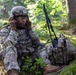 A U.S. Army soldier in full combat gear is seated at the base of a tree in a forested environment, using a radio handset for communication. The soldier is wearing MultiCam (OCP) camouflage uniform, a tactical helmet, and protective eyewear, with face paint applied. A tactical radio with an antenna is visible propped against a backpack beside the soldier.