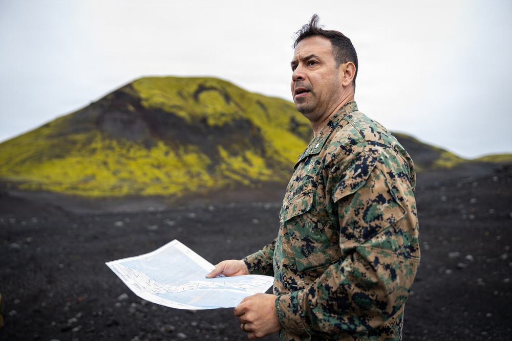 A U.S. Marine in MARPAT digital camouflage uniform holds a printed map or document while standing in a volcanic landscape with black terrain and moss-covered hills in the background. The individual appears to be conducting terrain reconnaissance or a briefing outdoors. The setting suggests a northern or subarctic environment, possibly Iceland or a similar volcanic region.