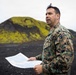 A U.S. Marine in MARPAT digital camouflage uniform holds a printed map or document while standing in a volcanic landscape with black terrain and moss-covered hills in the background. The individual appears to be conducting terrain reconnaissance or a briefing outdoors. The setting suggests a northern or subarctic environment, possibly Iceland or a similar volcanic region.