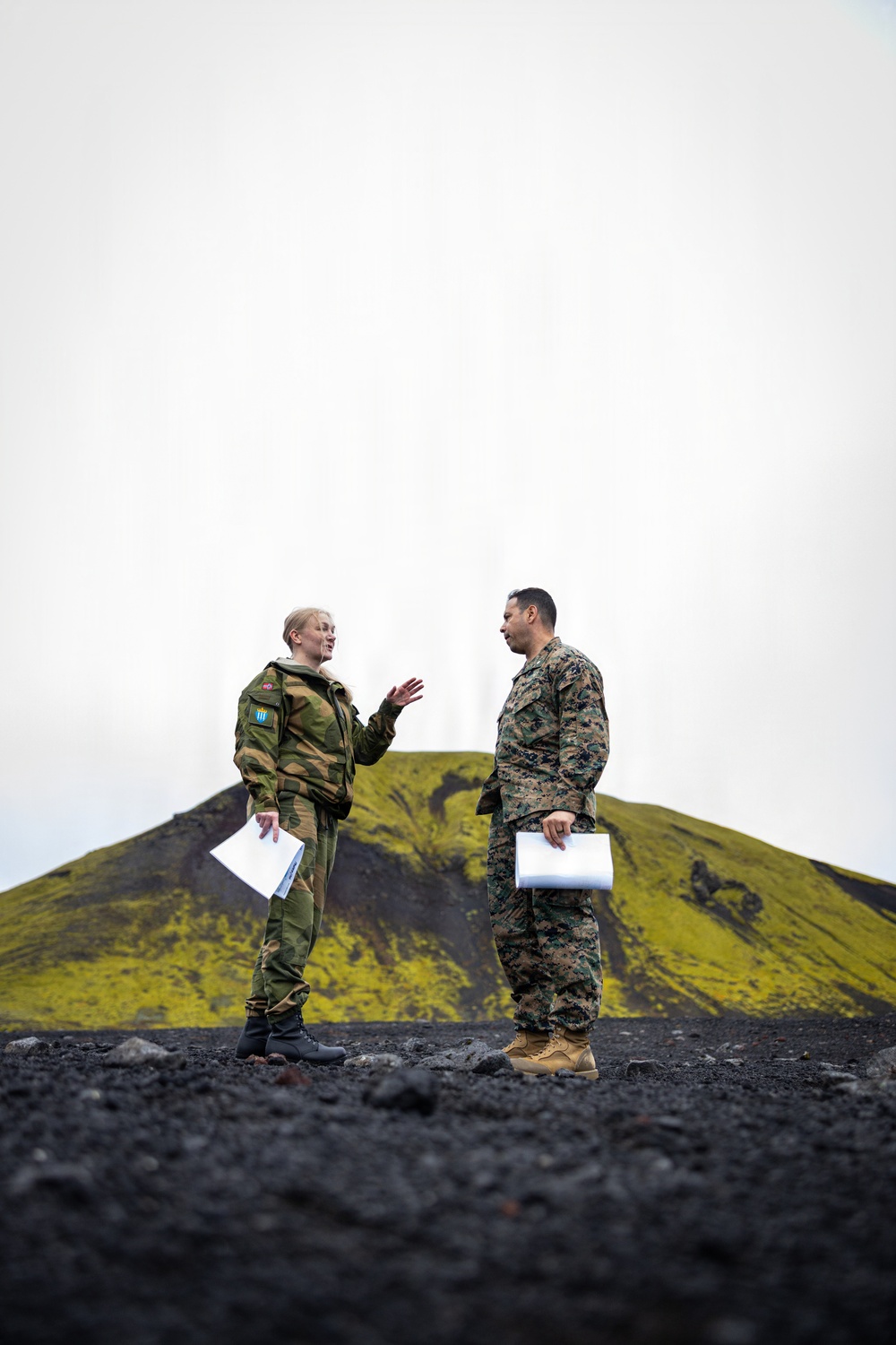 Two military personnel in different camouflage uniforms are engaged in conversation on a black volcanic terrain, with a moss-covered volcanic cone visible in the background. The individual on the left wears a distinctive woodland camouflage pattern with a shoulder patch featuring blue and yellow colors, while the individual on the right wears a MARPAT (US Marine Corps digital) camouflage uniform. Both are holding white documents or papers.