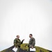 Two military personnel in different camouflage uniforms are engaged in conversation on a black volcanic terrain, with a moss-covered volcanic cone visible in the background. The individual on the left wears a distinctive woodland camouflage pattern with a shoulder patch featuring blue and yellow colors, while the individual on the right wears a MARPAT (US Marine Corps digital) camouflage uniform. Both are holding white documents or papers.