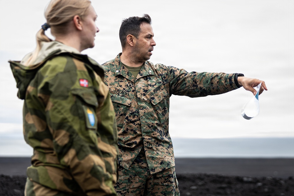 Two military personnel are engaged in what appears to be a briefing or reconnaissance discussion outdoors on dark volcanic terrain under an overcast sky. A U.S. Marine, identified by name tape as 'PALACIO' and wearing MARPAT digital camouflage, is pointing and gesturing while holding what appears to be a map or document. The second individual wears a Norwegian Armed Forces uniform, identifiable by the Norwegian flag patch and a blue/yellow shoulder insignia.