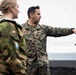 Two military personnel are engaged in what appears to be a briefing or reconnaissance discussion outdoors on dark volcanic terrain under an overcast sky. A U.S. Marine, identified by name tape as 'PALACIO' and wearing MARPAT digital camouflage, is pointing and gesturing while holding what appears to be a map or document. The second individual wears a Norwegian Armed Forces uniform, identifiable by the Norwegian flag patch and a blue/yellow shoulder insignia.