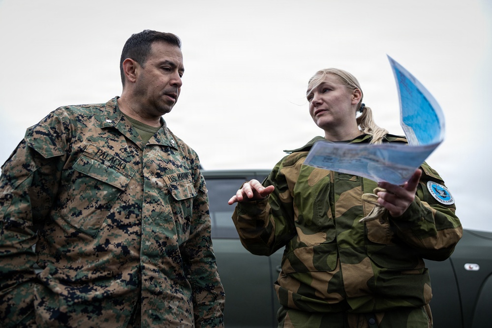 Two military personnel in different camouflage uniforms are engaged in a briefing or planning discussion outdoors. The individual on the left wears U.S. Marines digital camouflage (MARPAT) with a name tape reading 'PALACIO' and 'U.S. MARINES', while the individual on the right wears a woodland camouflage uniform with a circular patch and holds what appears to be a map or document. They appear to be conducting a joint military coordination activity near a military vehicle.