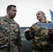Two military personnel in different camouflage uniforms are engaged in a briefing or planning discussion outdoors. The individual on the left wears U.S. Marines digital camouflage (MARPAT) with a name tape reading 'PALACIO' and 'U.S. MARINES', while the individual on the right wears a woodland camouflage uniform with a circular patch and holds what appears to be a map or document. They appear to be conducting a joint military coordination activity near a military vehicle.