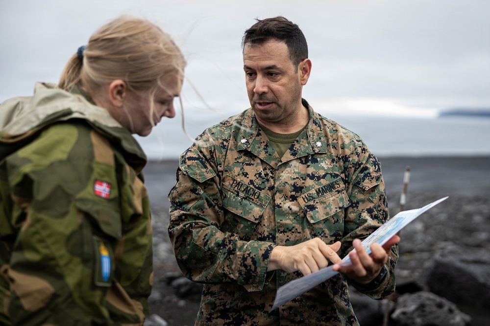 A U.S. Marine officer, identified by name tape as 'Palacio' and wearing the rank of Lieutenant Colonel (silver oak leaf), is reviewing a document or map with a Norwegian military personnel wearing a jacket bearing the Norwegian flag patch. The interaction appears to be a briefing or coordination meeting in an outdoor coastal or arctic environment. The setting suggests a multinational military exercise or operation.