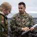 A U.S. Marine officer, identified by name tape as 'Palacio' and wearing the rank of Lieutenant Colonel (silver oak leaf), is reviewing a document or map with a Norwegian military personnel wearing a jacket bearing the Norwegian flag patch. The interaction appears to be a briefing or coordination meeting in an outdoor coastal or arctic environment. The setting suggests a multinational military exercise or operation.