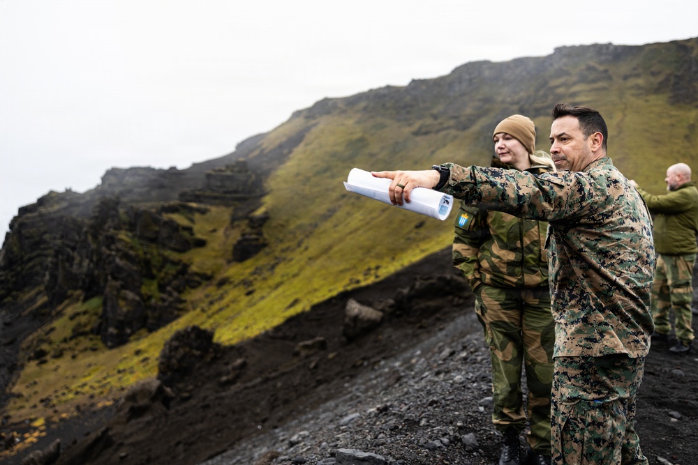 Two military personnel in camouflage uniforms stand on a rocky, moss-covered highland terrain, with one individual pointing into the distance while holding rolled documents. A third person is visible in the background. The setting appears to be a rugged volcanic or mountainous landscape under overcast skies.