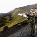 Two military personnel in camouflage uniforms stand on a rocky, moss-covered highland terrain, with one individual pointing into the distance while holding rolled documents. A third person is visible in the background. The setting appears to be a rugged volcanic or mountainous landscape under overcast skies.
