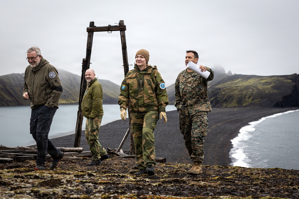 Four individuals are walking along a dark volcanic beach with dramatic mountainous terrain in the background. Two appear to be in military uniforms — one in US Marine Corps MARPAT camouflage and one in what appears to be Norwegian Armed Forces camouflage — while the other two wear civilian outdoor gear. The rightmost person is gesturing and appears to be briefing or directing the group.