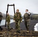 Four individuals are walking along a dark volcanic beach with dramatic mountainous terrain in the background. Two appear to be in military uniforms — one in US Marine Corps MARPAT camouflage and one in what appears to be Norwegian Armed Forces camouflage — while the other two wear civilian outdoor gear. The rightmost person is gesturing and appears to be briefing or directing the group.