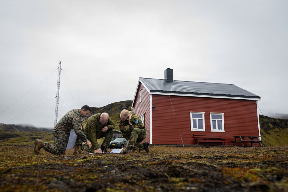 Three military personnel in camouflage uniforms are crouched on the ground examining a map or document outdoors. They are positioned in front of a red wooden building with a metal roof, set against a rugged, overcast landscape with green hills. The scene suggests a field planning or navigation exercise in a remote, likely Nordic or Arctic environment.