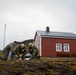 Three military personnel in camouflage uniforms are crouched on the ground examining a map or document outdoors. They are positioned in front of a red wooden building with a metal roof, set against a rugged, overcast landscape with green hills. The scene suggests a field planning or navigation exercise in a remote, likely Nordic or Arctic environment.