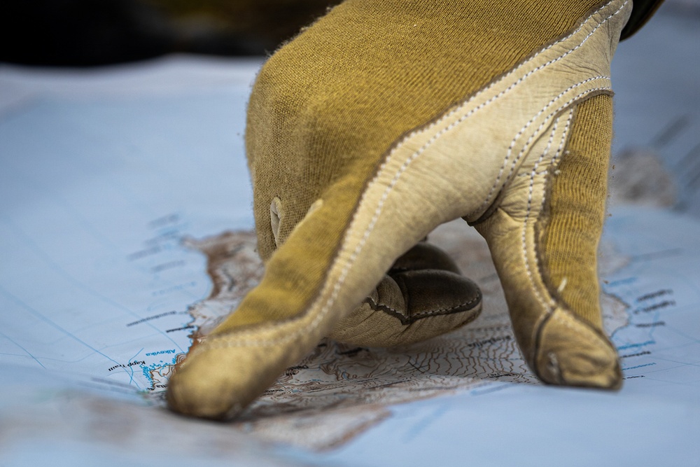 A gloved hand points to a location on a topographic map. The glove appears to be a tan/olive-colored tactical or work glove with leather reinforcement on the fingertips and palm. The image is a close-up shot with shallow depth of field, keeping the pointing finger and map in focus.