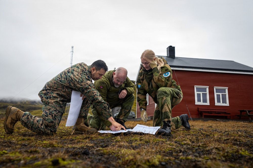 Three military personnel in different camouflage uniforms are crouched on sparse, mossy ground examining a map or document together. One individual wears US Marine Corps MARPAT digital camouflage, while the other two wear what appears to be Norwegian Armed Forces M98 camouflage pattern. They are positioned in front of a traditional red Scandinavian-style building in an overcast, cold-weather environment.