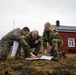 Three military personnel in different camouflage uniforms are crouched on sparse, mossy ground examining a map or document together. One individual wears US Marine Corps MARPAT digital camouflage, while the other two wear what appears to be Norwegian Armed Forces M98 camouflage pattern. They are positioned in front of a traditional red Scandinavian-style building in an overcast, cold-weather environment.