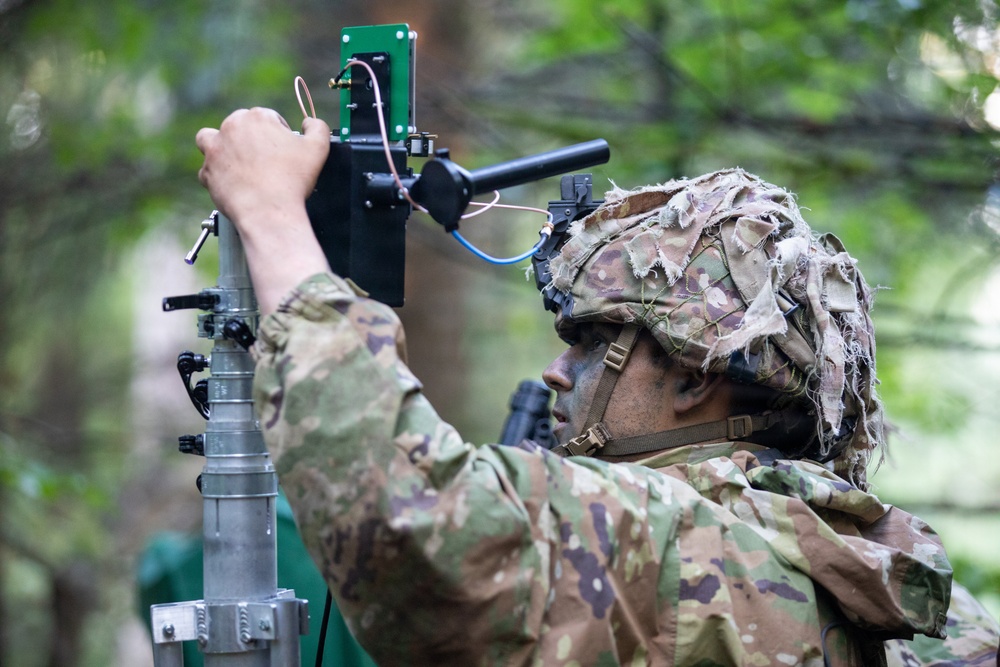 A soldier in full camouflage uniform and helmet with ghillie cover is adjusting or installing electronic equipment mounted on a telescoping mast or pole. The equipment appears to include a green circuit board, wiring, and a sensor or antenna assembly. The setting is a wooded outdoor environment, suggesting a field training exercise or operational deployment.