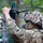 A soldier in full camouflage uniform and helmet with ghillie cover is adjusting or installing electronic equipment mounted on a telescoping mast or pole. The equipment appears to include a green circuit board, wiring, and a sensor or antenna assembly. The setting is a wooded outdoor environment, suggesting a field training exercise or operational deployment.