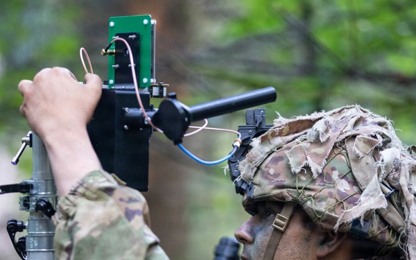 A soldier in full camouflage uniform and helmet with ghillie cover is adjusting or installing electronic equipment mounted on a telescoping mast or pole. The equipment appears to include a green circuit board, wiring, and a sensor or antenna assembly. The setting is a wooded outdoor environment, suggesting a field training exercise or operational deployment.