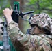 A soldier in full camouflage uniform and helmet with ghillie cover is adjusting or installing electronic equipment mounted on a telescoping mast or pole. The equipment appears to include a green circuit board, wiring, and a sensor or antenna assembly. The setting is a wooded outdoor environment, suggesting a field training exercise or operational deployment.