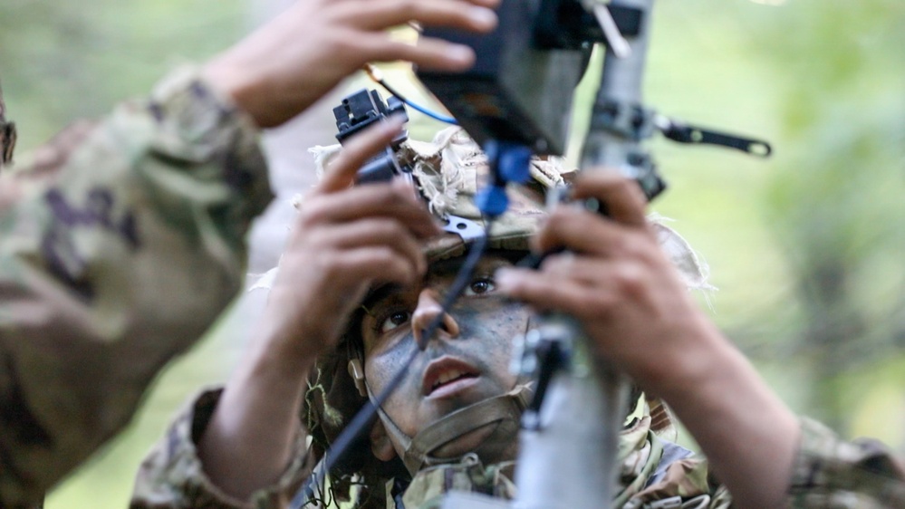 A soldier in camouflage uniform and face paint is closely examining or operating a piece of military optical or targeting equipment. The soldier appears to be looking through or adjusting a sighting device mounted on a stand or tripod. A second individual in camouflage is partially visible on the left side of the frame, also handling the equipment.