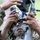 A soldier in camouflage uniform and face paint is closely examining or operating a piece of military optical or targeting equipment. The soldier appears to be looking through or adjusting a sighting device mounted on a stand or tripod. A second individual in camouflage is partially visible on the left side of the frame, also handling the equipment.