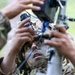 A soldier in camouflage uniform and face paint is closely examining or operating a piece of military optical or targeting equipment. The soldier appears to be looking through or adjusting a sighting device mounted on a stand or tripod. A second individual in camouflage is partially visible on the left side of the frame, also handling the equipment.