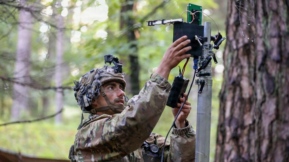 A soldier in multicam camouflage uniform and helmet is installing or adjusting an electronic device mounted on a pole in a forested environment. The device appears to be a sensor, antenna, or surveillance equipment with visible circuit boards and wiring. The setting suggests a field exercise or operational deployment in a woodland area.