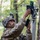 A soldier in multicam camouflage uniform and helmet is installing or adjusting an electronic device mounted on a pole in a forested environment. The device appears to be a sensor, antenna, or surveillance equipment with visible circuit boards and wiring. The setting suggests a field exercise or operational deployment in a woodland area.