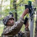 A soldier in multicam camouflage uniform and helmet is installing or adjusting an electronic device mounted on a pole in a forested environment. The device appears to be a sensor, antenna, or surveillance equipment with visible circuit boards and wiring. The setting suggests a field exercise or operational deployment in a woodland area.