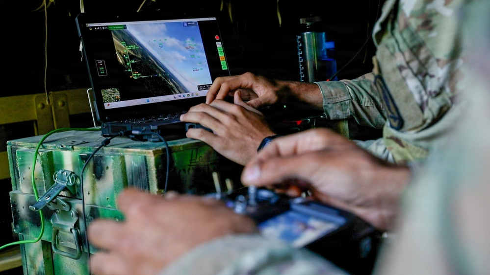 Military personnel in camouflage uniforms are operating a drone or UAV ground control station. The setup includes a laptop displaying aerial/flight telemetry data and a handheld radio controller, mounted on a military-grade equipment case. The scene appears to be an active drone operation or training exercise.