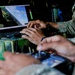 Military personnel in camouflage uniforms are operating a drone or UAV ground control station. The setup includes a laptop displaying aerial/flight telemetry data and a handheld radio controller, mounted on a military-grade equipment case. The scene appears to be an active drone operation or training exercise.