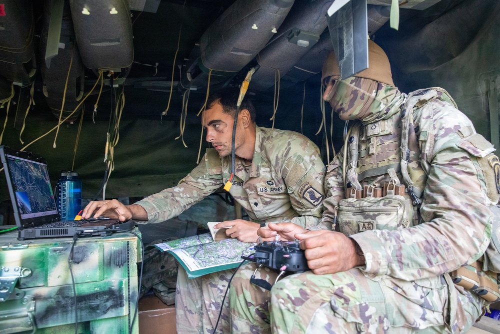 Two U.S. Army soldiers are working inside what appears to be a military command vehicle or tactical operations center. The soldier on the left is operating a ruggedized laptop displaying what appears to be a map or terrain visualization, while the soldier on the right holds a handheld device and references a paper map. Both are wearing Operational Camouflage Pattern (OCP) uniforms in what appears to be a field training or operational environment.