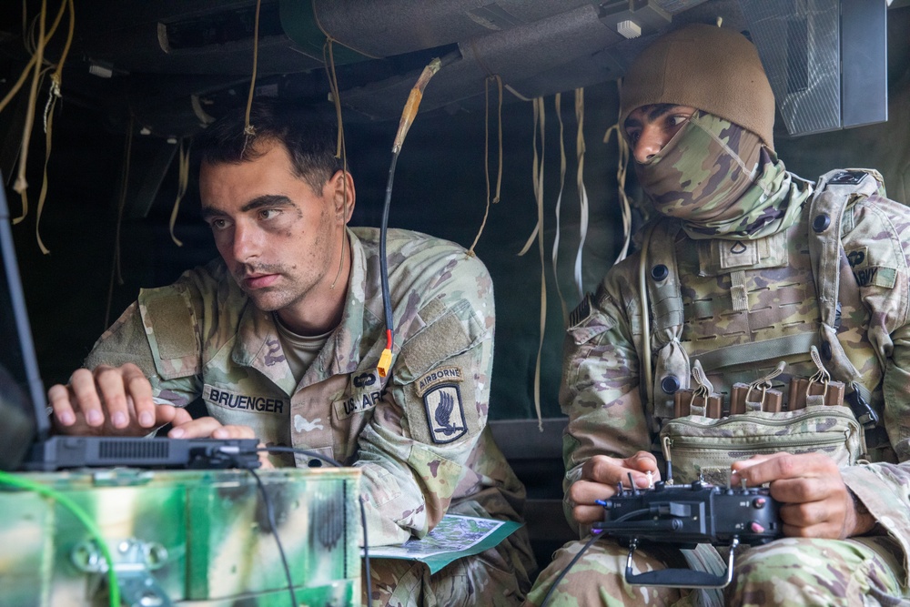 Two U.S. Army soldiers are seated inside a military vehicle, engaged in what appears to be drone or UAS (Unmanned Aerial System) operations. The soldier on the left, identified by name tape as 'BRUENGER,' is operating a laptop or tactical computer, while the soldier on the right holds a remote control device. Both wear OCP (Operational Camouflage Pattern) uniforms, with the left soldier displaying a 173rd Airborne Brigade patch.