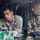 Two U.S. Army soldiers are seated inside a military vehicle, engaged in what appears to be drone or UAS (Unmanned Aerial System) operations. The soldier on the left, identified by name tape as 'BRUENGER,' is operating a laptop or tactical computer, while the soldier on the right holds a remote control device. Both wear OCP (Operational Camouflage Pattern) uniforms, with the left soldier displaying a 173rd Airborne Brigade patch.