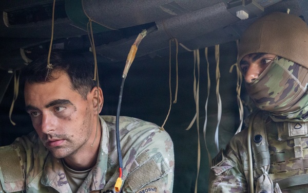 Two U.S. Army soldiers are seated inside a military vehicle, engaged in what appears to be drone or UAS (Unmanned Aerial System) operations. The soldier on the left, identified by name tape as 'BRUENGER,' is operating a laptop or tactical computer, while the soldier on the right holds a remote control device. Both wear OCP (Operational Camouflage Pattern) uniforms, with the left soldier displaying a 173rd Airborne Brigade patch.