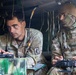 Two U.S. Army soldiers are seated inside a military vehicle, engaged in what appears to be drone or UAS (Unmanned Aerial System) operations. The soldier on the left, identified by name tape as 'BRUENGER,' is operating a laptop or tactical computer, while the soldier on the right holds a remote control device. Both wear OCP (Operational Camouflage Pattern) uniforms, with the left soldier displaying a 173rd Airborne Brigade patch.