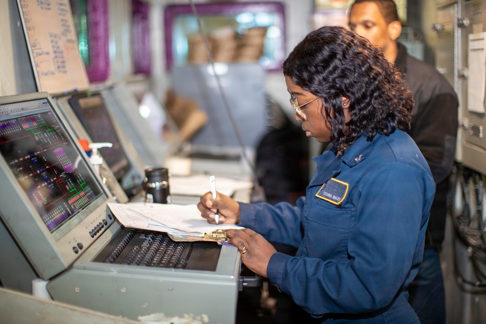 A uniformed military or naval personnel member, identified by a name tag reading 'KESHA DALEY', is working at a control station with a monitor displaying what appears to be a systems diagram or schematic. She is writing notes in a binder/clipboard while reviewing the screen. A second individual is visible in the background in a darker uniform.