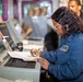 A uniformed military or naval personnel member, identified by a name tag reading 'KESHA DALEY', is working at a control station with a monitor displaying what appears to be a systems diagram or schematic. She is writing notes in a binder/clipboard while reviewing the screen. A second individual is visible in the background in a darker uniform.