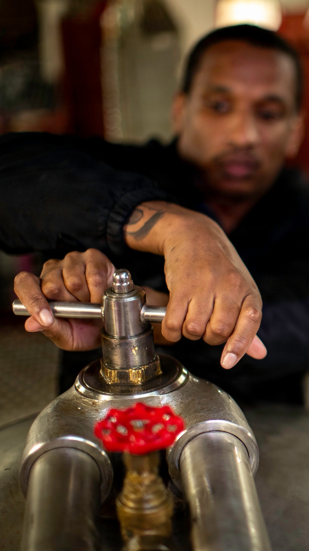 A man with a tattoo on his wrist is operating a metal valve or fitting on what appears to be a pressurized cylinder or industrial pipe assembly. He is using both hands to turn a T-bar handle on top of the fitting. A red wheel valve is also visible on the lower portion of the equipment.