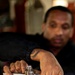 A man with a tattoo on his wrist is operating a metal valve or fitting on what appears to be a pressurized cylinder or industrial pipe assembly. He is using both hands to turn a T-bar handle on top of the fitting. A red wheel valve is also visible on the lower portion of the equipment.