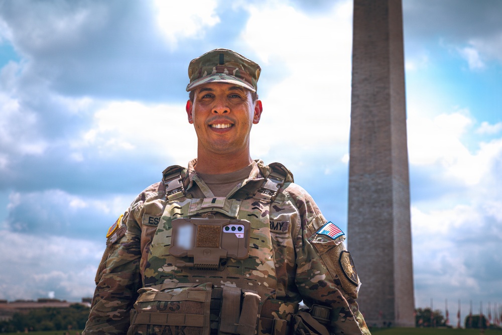 A U.S. Army soldier in full Operational Camouflage Pattern (OCP) uniform and body armor is posing and smiling in front of the Washington Monument in Washington, D.C. The soldier is wearing a patrol cap, plate carrier vest with attached equipment, and has visible unit patches and a U.S. flag patch on the right sleeve. The image appears to be an official or semi-official military portrait taken on the National Mall.
