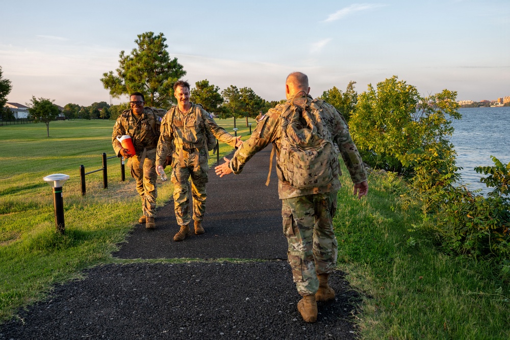 Three military personnel in camouflage uniforms are walking along a paved path near a waterfront area during golden hour lighting. One soldier facing away from the camera is extending his hand to greet an approaching soldier who is smiling. The setting appears to be a military base or park with manicured grass, trees, and a body of water visible in the background.