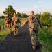 Three military personnel in camouflage uniforms are walking along a paved path near a waterfront area during golden hour lighting. One soldier facing away from the camera is extending his hand to greet an approaching soldier who is smiling. The setting appears to be a military base or park with manicured grass, trees, and a body of water visible in the background.