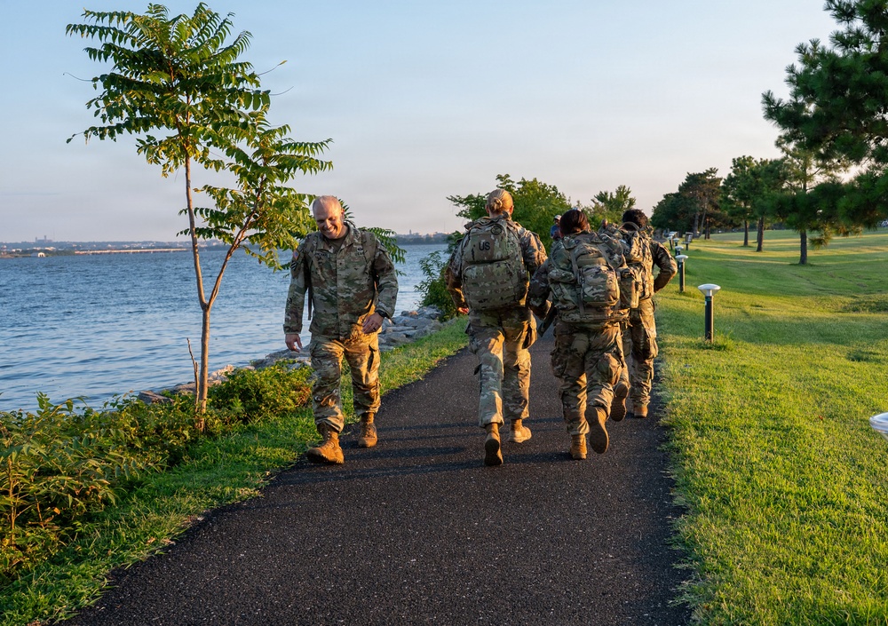 A group of military personnel in camouflage uniforms and tactical gear are walking along a paved waterfront path during early morning or evening hours. They are carrying rucksacks and appear to be conducting a ruck march or physical training exercise. The setting is a well-maintained park or military installation alongside a large body of water.