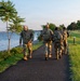 A group of military personnel in camouflage uniforms and tactical gear are walking along a paved waterfront path during early morning or evening hours. They are carrying rucksacks and appear to be conducting a ruck march or physical training exercise. The setting is a well-maintained park or military installation alongside a large body of water.