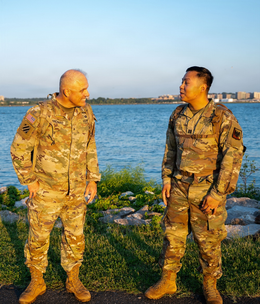 Two U.S. Army soldiers in Operational Camouflage Pattern (OCP) uniforms are standing near a waterfront, engaged in conversation. The soldier on the left displays a single star insignia indicating a Brigadier General rank, with a name tape reading 'ANCHARD' and a 3rd Infantry Division patch. The soldier on the right wears Captain rank insignia (two vertical bars) with a name tape partially reading 'NN' and a different unit patch.
