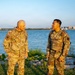 Two U.S. Army soldiers in Operational Camouflage Pattern (OCP) uniforms are standing near a waterfront, engaged in conversation. The soldier on the left displays a single star insignia indicating a Brigadier General rank, with a name tape reading 'ANCHARD' and a 3rd Infantry Division patch. The soldier on the right wears Captain rank insignia (two vertical bars) with a name tape partially reading 'NN' and a different unit patch.
